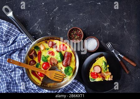 Frittata mit Würstchen, Brokkoli, Kirschtomaten, geschmolzenem Käse in einem Skillet auf einem Betontisch und einem Teil auf einem Teller, horizontale Ansicht von oben Stockfoto