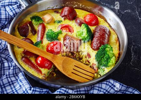 Frittata mit Würstchen, Brokkoli, Kirschtomaten, geschmolzenem Käse in einem Skillet mit Spachtel auf einem Betontisch, horizontale Ansicht von oben, Nahaufnahme Stockfoto