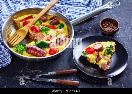 Nahaufnahme von Omelette mit Würstchen, Brokkoli, Kirschtomaten, geschmolzenem Käse in einem Skillet auf einem Betontisch und einem Teil auf einem Teller, horizontale Ansicht Stockfoto