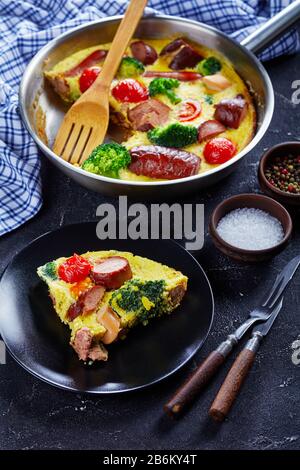 Frittata mit Würstchen, Brokkoli, Kirschtomaten, geschmolzenem Käse in einem Skillet auf einem Betontisch und einem Teil auf einem Teller, vertikale Ansicht von oben, Stockfoto