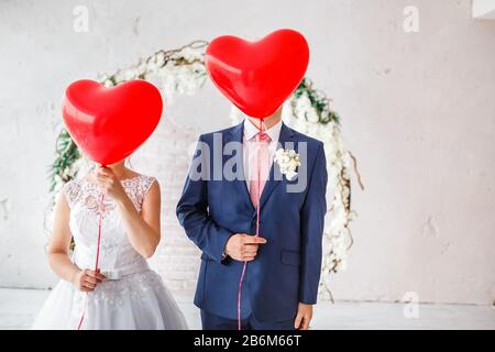 Fröhliche Bräutigam und Brautpaar bedeckten ihre Gesichter mit roten Luftballons auf der Hochzeitsfeier in Innenräumen Stockfoto