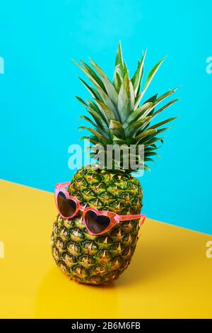 Ananas mit einer Paar rosafarbenen, herzförmigen Sonnenbrillen auf blauem und gelbem Hintergrund Stockfoto