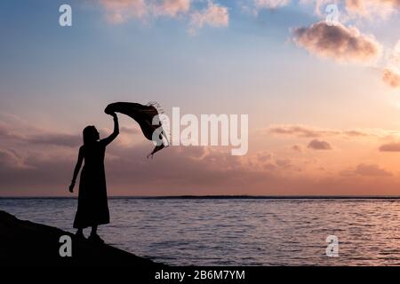 Silhouette asian Woman erholt sich mit hochgezogenen Lappen auf tropischem Meer mit farbenfrohem Himmel Stockfoto