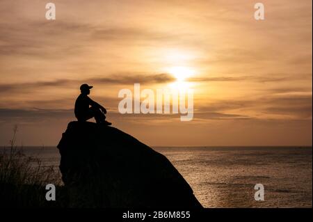 Silhouette man sitzt auf einem großen Felsen mit Sightseeing bei Sonnenuntergang im tropischen Meer Stockfoto