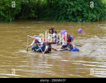 Vier Männer in einem schicken Kleid, die während eines Floßrennens ein hausgemachte Floß auf dem Fluss Severn in Shrewsbury paddeln. Stockfoto