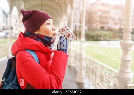 Junge Frau in der Jacke trinkt therapeutisches Mineralwasser aus einer Tasse eine natürliche heiße Quelle in Karlsbad zur Winterzeit, Tschechien Stockfoto