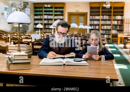 Niedliches kleines Mädchen verwendet die Anwendung auf dem Tablet, und ein älterer Mann liest ein Buch in der Bibliothek. Alte und neue Technologien, Papierbuch und eBook Stockfoto