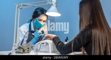 Eine Frau, die im Salon Maniküre meistt und dabei mit den Nägeln des Kunden auf blauem Hintergrund arbeitet. Kosmetologie und Nagelpflege Stockfoto