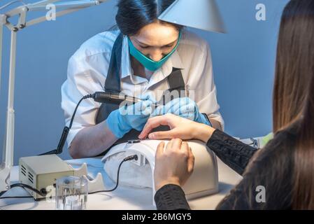 Eine Frau, die im Salon Maniküre meistt und dabei mit den Nägeln des Kunden auf blauem Hintergrund arbeitet. Kosmetologie und Nagelpflege Stockfoto