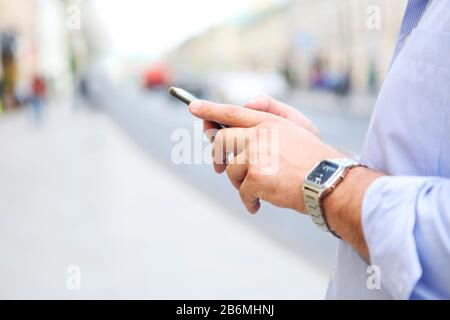 Seitenansicht von Crop männlich in blauem Hemd und Armbanduhr mit Smartphone beim stehen auf der City Street Stockfoto