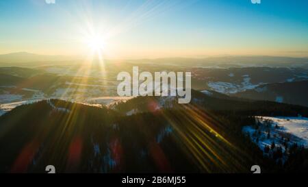 Luftbild sonnige schöne Landschaft von Winterhügeln und Bergen mit Walddickichten an einem frostigen Winterabend gegen einen blauen Himmel und die Umgebung Stockfoto