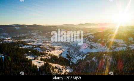 Luftbild sonnige schöne Landschaft von Winterhügeln und Bergen mit Walddickichten an einem frostigen Winterabend gegen einen blauen Himmel und die Umgebung Stockfoto