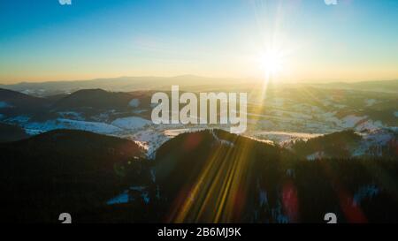 Luftbild sonnige schöne Landschaft von Winterhügeln und Bergen mit Walddickichten an einem frostigen Winterabend gegen einen blauen Himmel und die Umgebung Stockfoto