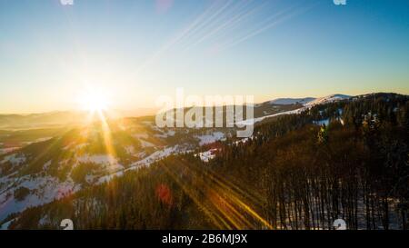 Luftbild sonnige schöne Landschaft von Winterhügeln und Bergen mit Walddickichten an einem frostigen Winterabend gegen einen blauen Himmel und die Umgebung Stockfoto