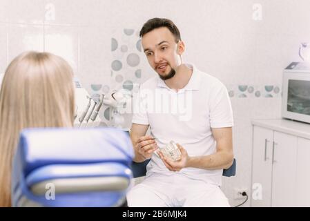 Der Zahnarzt erzählt am Beispiel eines künstlichen Kiefers, wie man um Spangen kümmert. Eine Frau, eine Zahnärztin, sitzt in einem Zahnstuhl und hört sich eine Arztgeschichte über Mundhygiene an. Stockfoto