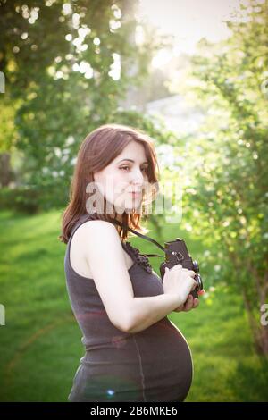 Seitenansicht der charmanten jungen schwangeren Frau im lässigen Kleid aufnehmen mit Fotokamera im grünen Park am sonnigen Sommertag Stockfoto