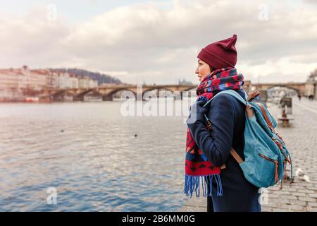 Junge Frau Touristin im Mantel mit Rucksack in der Nähe der Brücke über die Moldau in Prag Stockfoto