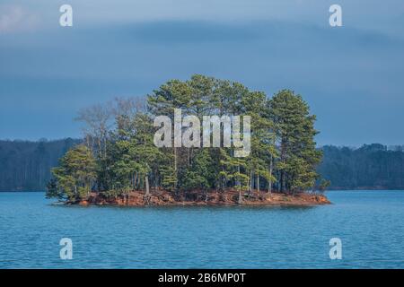Eine Insel inmitten des Lake Lanier Georgia mit dem Sonnenlicht, das durch den stürmischen Himmel schält und die Bäume mit dem Wald im Rücken hervorhebt Stockfoto