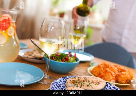 Holztisch serviert mit köstlichen Gerichten und Getränken für die Heimparty, mit Getreide, der nicht erkennbar ist, und Wein im Hintergrund auf Glas gießt Stockfoto