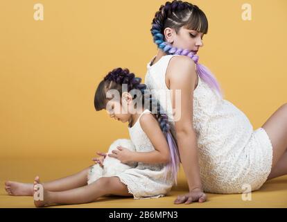 Positiv lachende Mutter und Tochter mit den gleichen Frisuren und Kleidern, die im Studio auf dem Boden sitzen und mit einem schwangeren Bauch und Ball posieren. Stockfoto