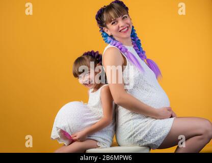 Positiv lachende Mutter und Tochter mit den gleichen Frisuren und Kleidern, die im Studio auf dem Boden sitzen und mit einem schwangeren Bauch und Ball posieren. Stockfoto