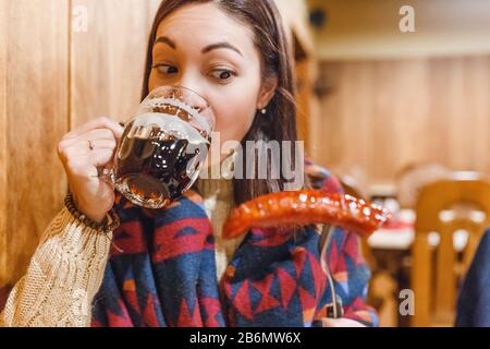 Eine Frau in einem Pub trinkt Kraftbier aus einem Vintage-Becher und Snacks eine Schweinewurst Stockfoto