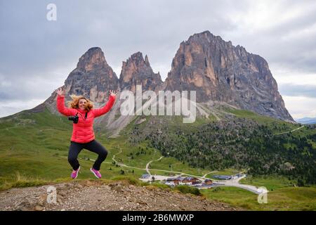 Fröhliche Fotografin, die mit ihren Armen in der Luft sprang und eine leuchtend rote Jacke trug, vor dem Sassolungo (Saslonch, Langkofel) Berg, in den Dolmen, Stockfoto