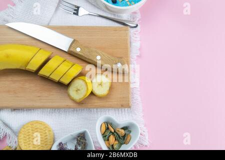 Geschnittene Bananen auf einem Holztablett, neben der Salatschüssel sind Mandelsandnüsse und Kürbiskerne Stockfoto