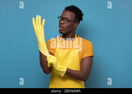 Der junge afroamerikanische Mann, der mit Handschuhen über isoliertem gelbem Hintergrund reinigt, ist bereit für das Waschen und andere Hausaufgaben. Studio Sho Stockfoto