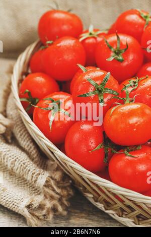 Frische rote Tomaten in einem Korbkorb auf einem alten Holztisch. Reife und saftige Kirschtomaten mit Tropfen Feuchtigkeit, grauer Holztisch, um ein Tuch Stockfoto