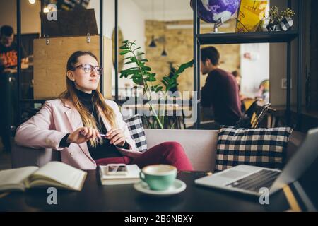 Schöne kaukasische Frau träumt von etwas, während sie mit dem Computer im modernen Café sitzt. Freiberuflerin denkt während der Arbeit über neue Ideen nach Stockfoto