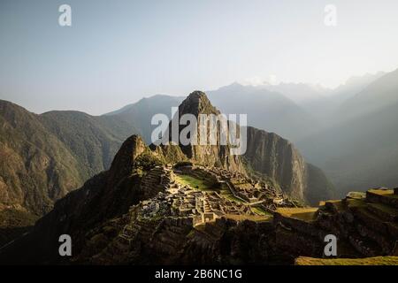 Weltberühmte Inka-Stadt Machu Picchu bei Sonnenaufgang mit gelben Lichtstrahlen und nebligen Wolken (ohne Besucher) (Aguas Calientes, Peru) Stockfoto