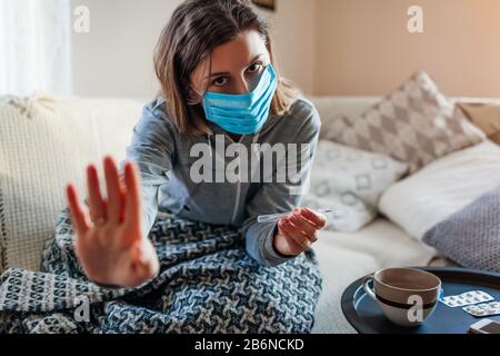 Erkrankte Frau überprüft Thermometer mit Fieber. Mädchen trägt eine Schutzmaske mit kalter Grippe zu Hause zeigt Stoppschild. Stockfoto