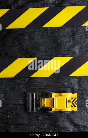 Spielzeugbagger gelb auf schwarzem texturalem Hintergrund. Band Fechten schwarz gelb vor dem Hintergrund des KONZEPTS. Vertikaler Rahmen Stockfoto