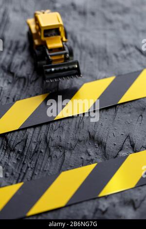 Spielzeugbagger gelb auf schwarzem texturalem Hintergrund. Band Fechten schwarz gelb vor dem Hintergrund des KONZEPTS. Vertikaler Rahmen Stockfoto