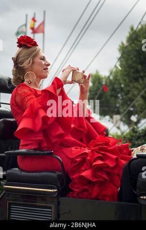 Dame, die ein sevillian Kostüm trägt, auf einer Pferdekutsche sitzt und mit ihrem Handy Fotos im Feria de Abril Enclosure in Sevilla, Spanien macht Stockfoto