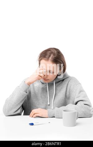 Kranke junge Frau, die mit Digitalthermometer und Becher am Tisch sitzt und Tränen wischt, während sie wegen Krankheit weint Stockfoto