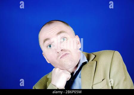Ein verwirrter oder überraschter junger Mann mit blauen Augen hat seine Faust unter sein Kinn gelegt. Emotionen Gesichtsausdruck. Stockfoto