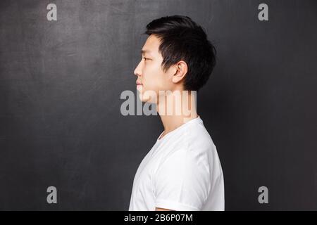 Profilportrait des jungen asiatischen männlichen Modells in weißem T-Shirt lächelnd beiläufig stehend und links über schwarzem Hintergrund des Studios, einsehend Stockfoto