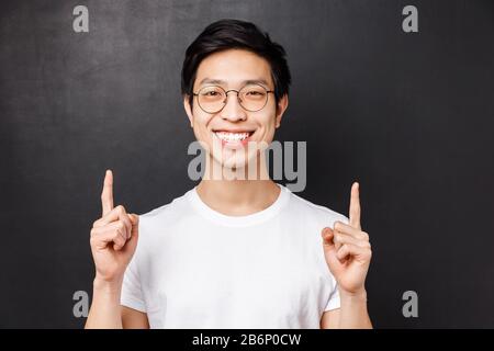 Nahporträt des fröhlichen, fröhlichen asiatischen jungen männlichen Studenten, der über seine Erfahrung im Auslandsstudium spricht und bei der Präsentation mit den Fingern nach oben zeigt Stockfoto