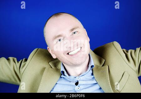Porträt des jungen Mannes mit schneeweißem Lächeln. Kurze Haare, cremefarbener Mantel und blauer Hintergrund. Emotionen und Lächeln Stockfoto