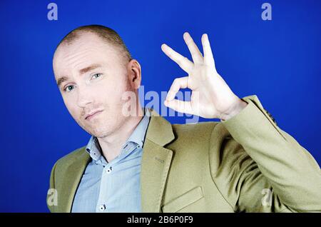 Ein selbstbewusster junger Mann in einem cremefarbenen Mantel zeigt Okeys Geste und blickt auf die Kamera. Gesturing und Schilder Konzept Stockfoto