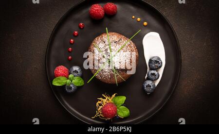 Schokoladenmuffins mit Himbeeren und Blaubeeren auf schwarzem Teller mit Hintergrundbeleuchtung, Lebensmittelfoto in hellen Farben Stockfoto