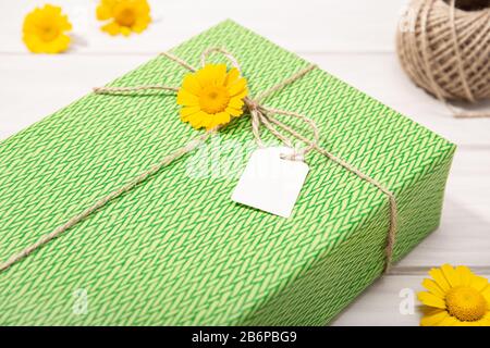 Grünes Geschenk oder Geschenkbox mit leerem Etikett und Blumen in Gänseblümchen auf dem Tisch. Verspotten Sie sich Stockfoto