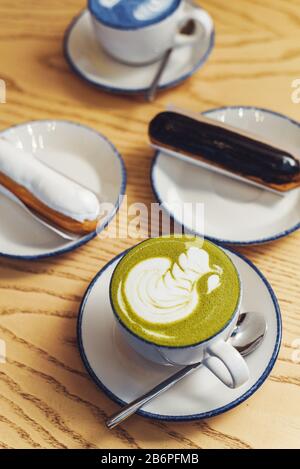 Blaue und grüne Matcha in Gläsern auf einem Holztisch in einem Café neben einem süßen Dessert. Eklairs mit Schokoladenganache und Vereisung mit verschiedenen Belägen. Stockfoto