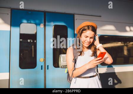 Eisenbahnthema. Schöne junge Frau mit Rucksack benutzt das Telefon, während sie in der Nähe des Bahnsteigs auf dem Bahnsteig steht. Günstige Reise Stockfoto