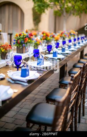 Tisch für eine Hochzeit auf der Terrasse mit orangefarbenen und gelben Blumen, blauen und kristallklaren Windgläsern auf einem rustikalen Tisch in einem Innenhof im Missionsstil Stockfoto