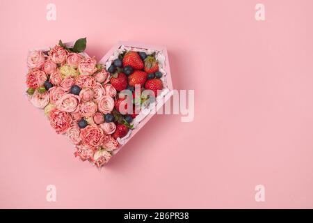 Holzkiste in Herzform mit Rosen und reifen Erdbeeren auf pinkfarbenem Hintergrund. Stockfoto