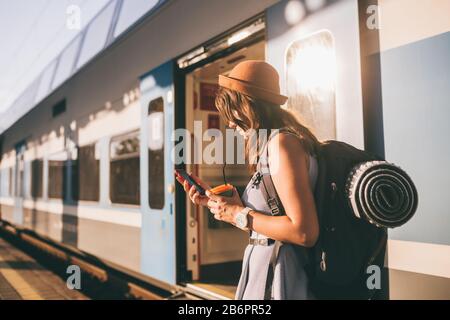 Eisenbahnthema. Schöne junge Frau mit Rucksack benutzt das Telefon, während sie in der Nähe des Bahnsteigs auf dem Bahnsteig steht. Günstige Reise Stockfoto