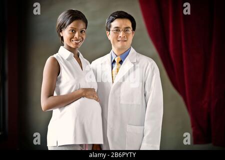Porträt einer schwangeren jungen Frau, die mit der Hand auf dem Bauch neben einem männlichen Arzt steht. Stockfoto